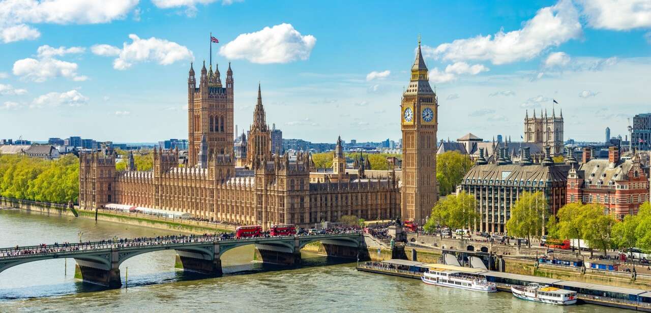 The Palace of Westminster and Big Ben in London, viewed from across the River Thames on a sunny day with Westminster Bridge and red double-decker buses in the foreground.