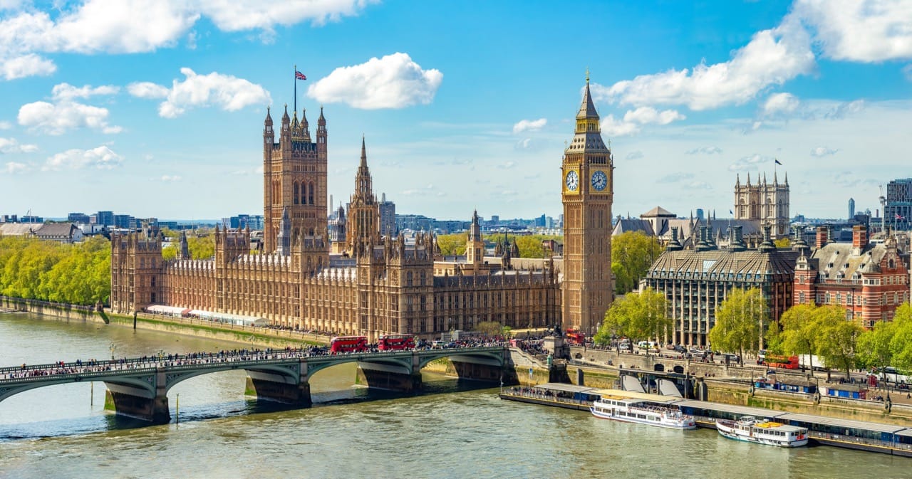 The Palace of Westminster and Big Ben in London, viewed from across the River Thames on a sunny day with Westminster Bridge and red double-decker buses in the foreground.